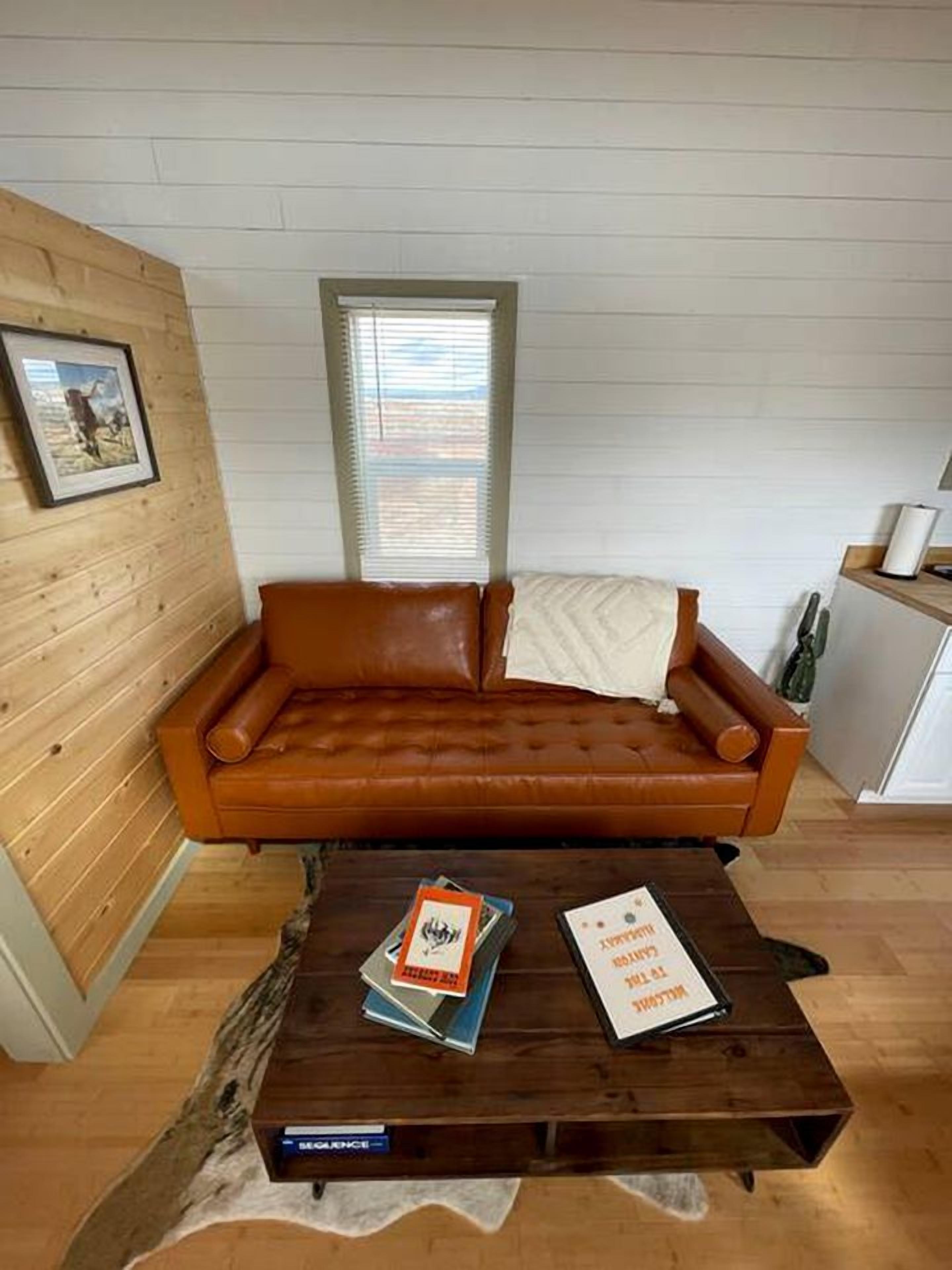 A room with a brown leather sofa, a wooden coffee table with books, a framed picture on the wall, and a window with blinds.