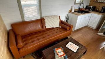 A cozy room with a brown leather sofa, a wooden coffee table, and a kitchen area with white cabinets and a microwave.