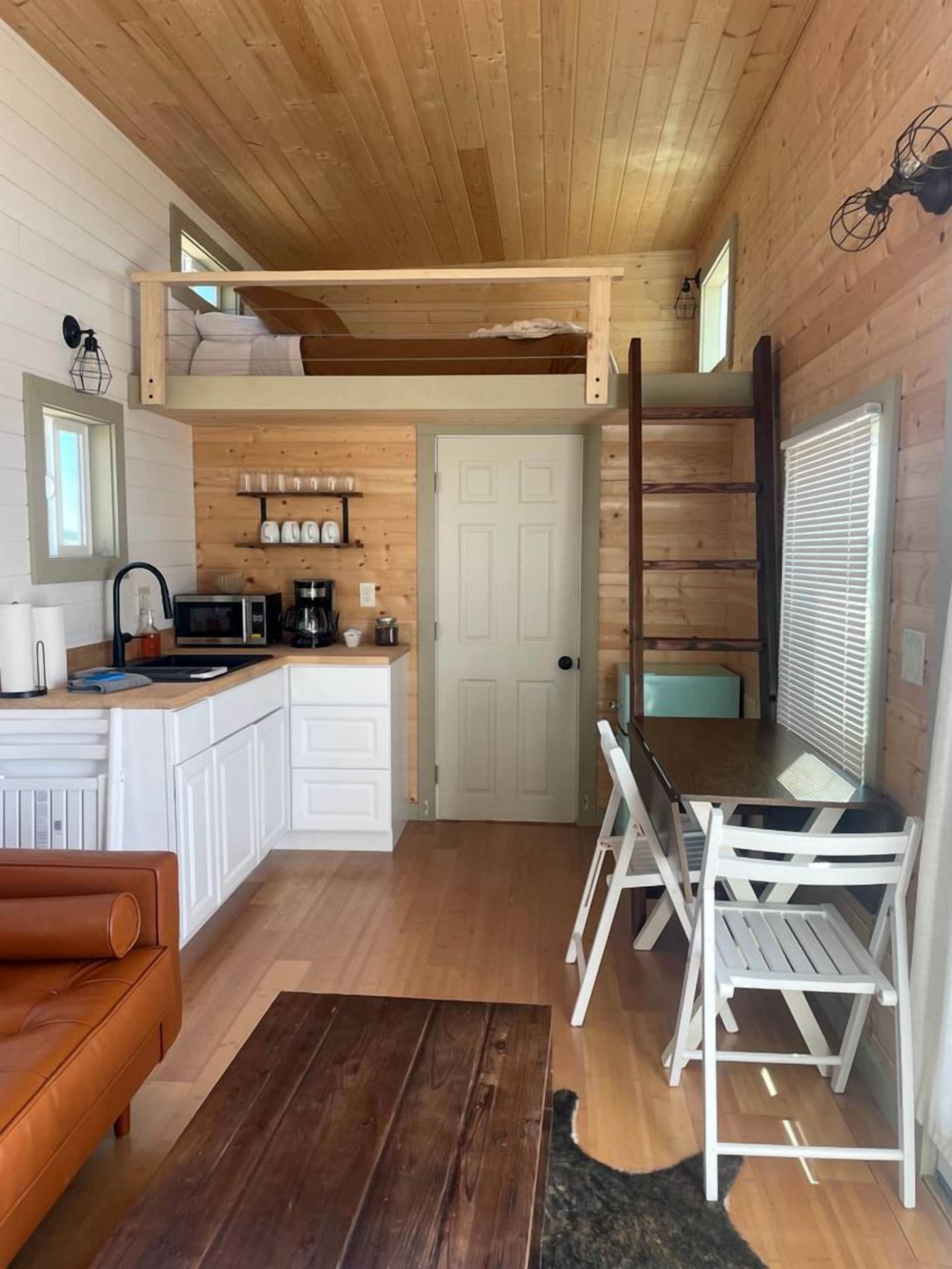 A compact kitchen with a wooden ceiling, white cabinets, a dining area with white chairs, and a brown leather sofa.