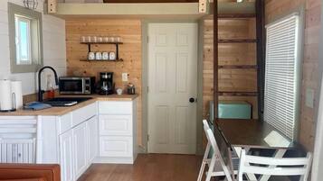 A compact kitchen with a wooden ceiling, white cabinets, a dining area with white chairs, and a brown leather sofa.