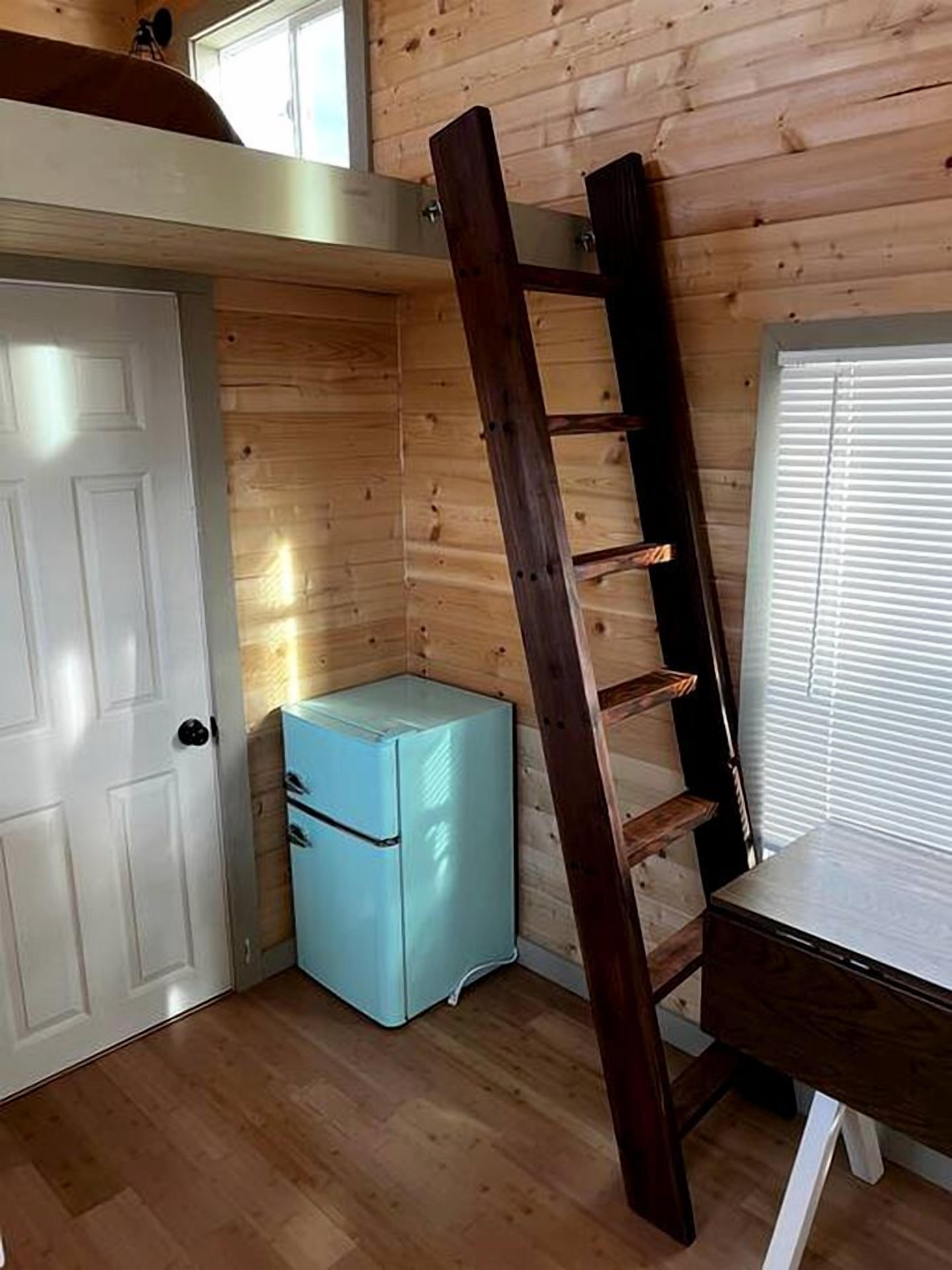 A room with a loft bed, a small blue refrigerator, a wooden ladder, a white door, and a window with blinds.