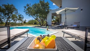 A poolside setting with a table holding a bowl of fruit, lounge chairs, and a clear view of the pool and surrounding trees. - Villa Karla with pool&sea view by Villas Guide (Banjol)