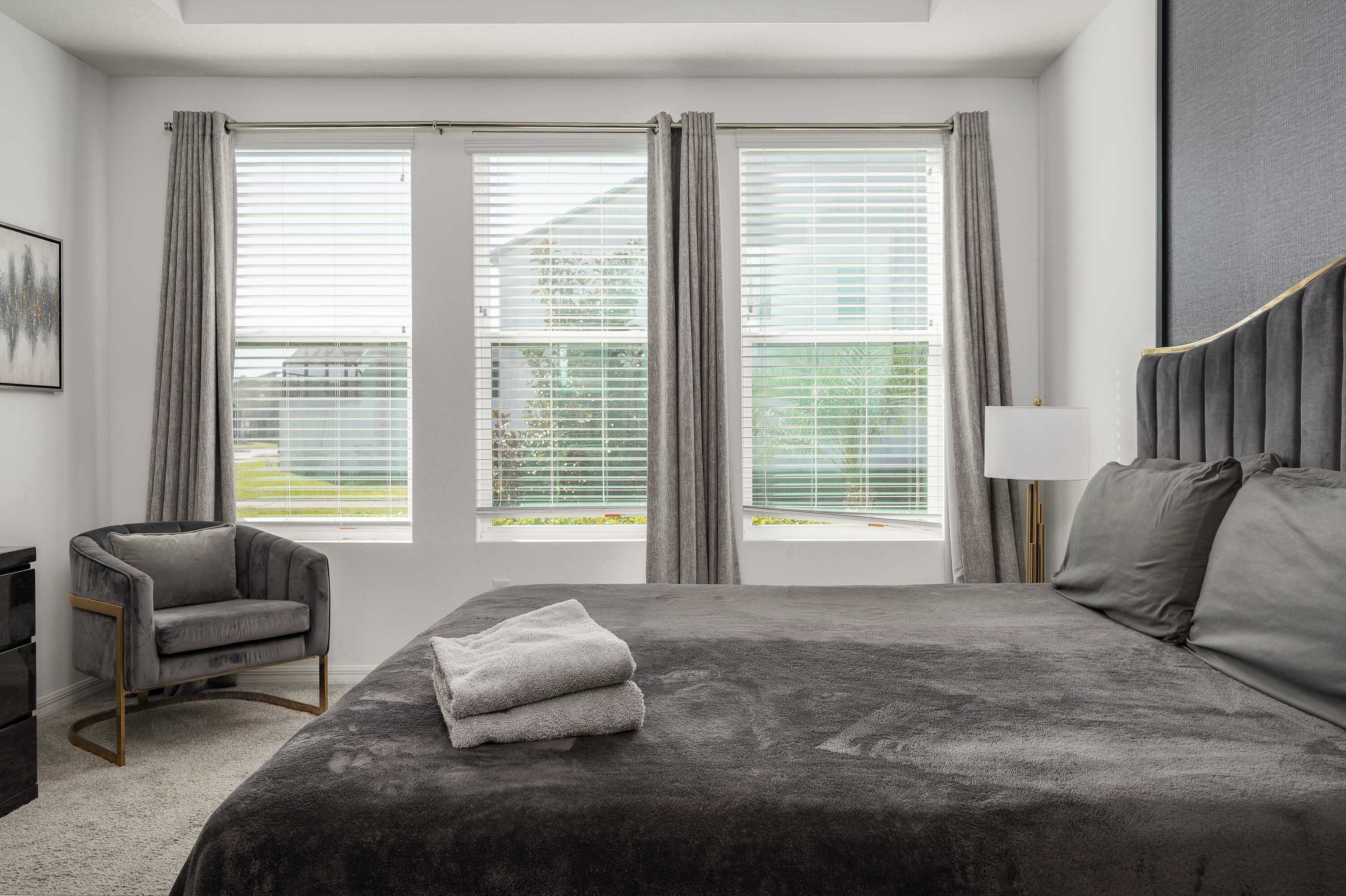 A bedroom with a large bed, two windows with curtains, a chair, and a bedside table.