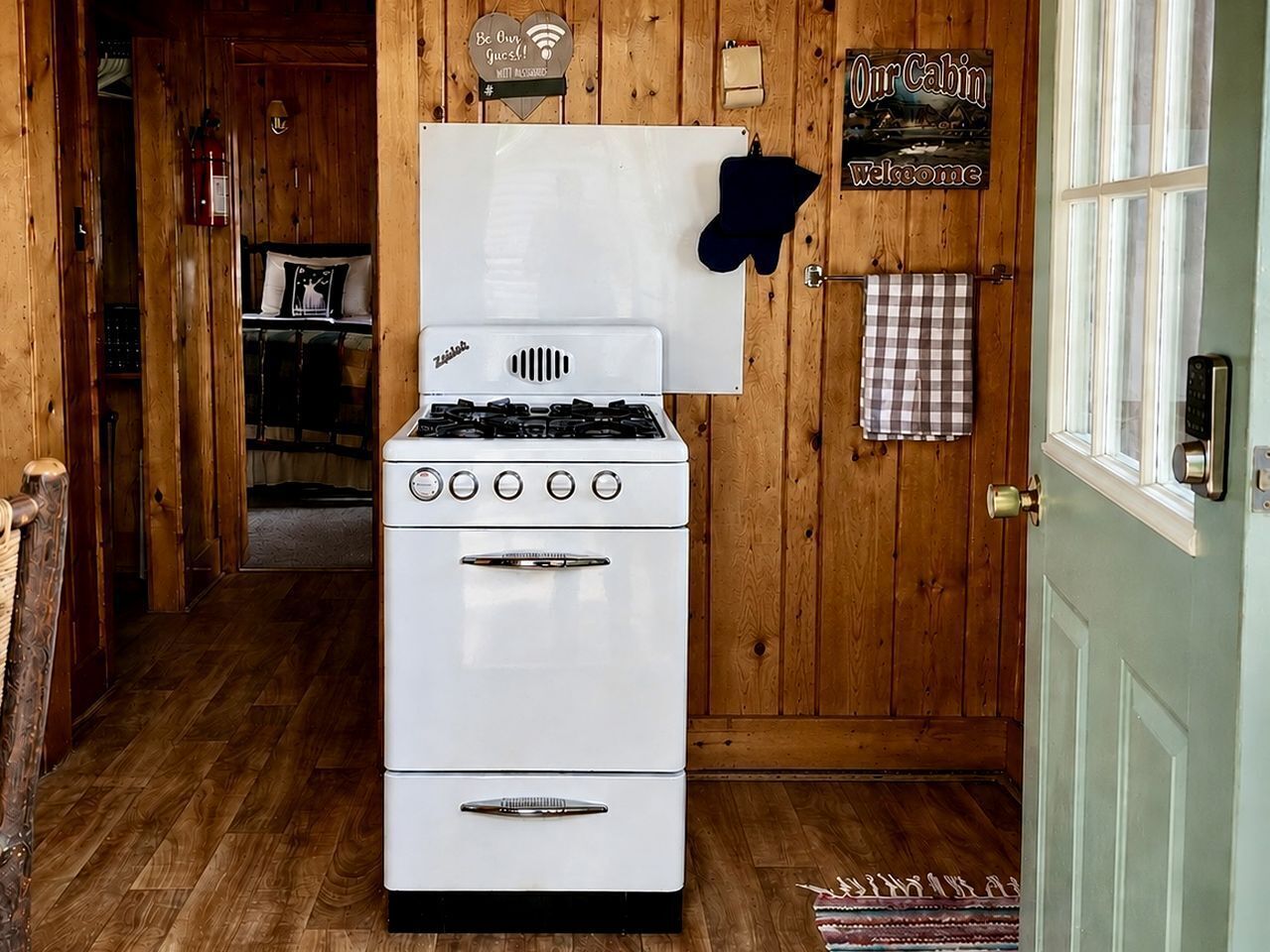 A white stove in a wooden cabin with a sign reading 'Our Cabin Welcome' and a black oven mitt on the wall.