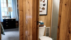 A wooden-paneled bathroom with a toilet, a framed animal illustration, and a window with curtains.