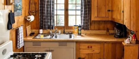 A cabin-style kitchen with wooden walls, a stove, a ceiling fan, and a window with curtains.