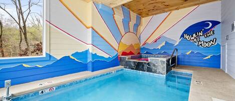 An indoor swimming pool with a mural of a sunrise and mountains, a hot tub, and a window with a view of trees.