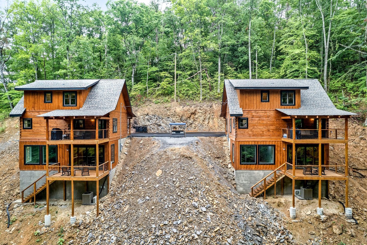 Two wooden cabins with balconies, set in a forested area with a rocky ground.