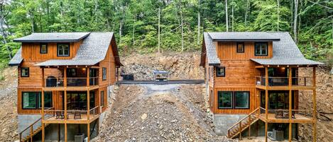 Two wooden cabins with balconies, set in a forested area with a rocky ground.