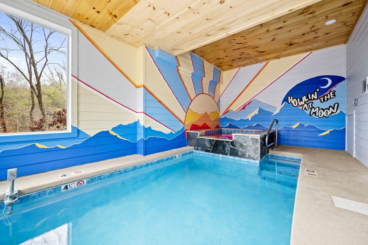 An indoor swimming pool with a mural of a sunrise and mountains, a hot tub, and a window with a view of trees.