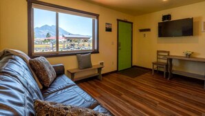 Una habitación con piso de madera, una puerta verde, un sofá, un banco, un televisor y una ventana con vista a la montaña.
