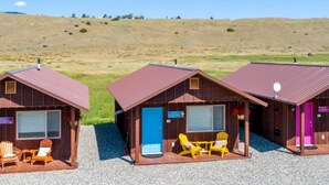 Tres cabañas de madera con suelo de grava, cada una con una puerta, ventana y una silla afuera.