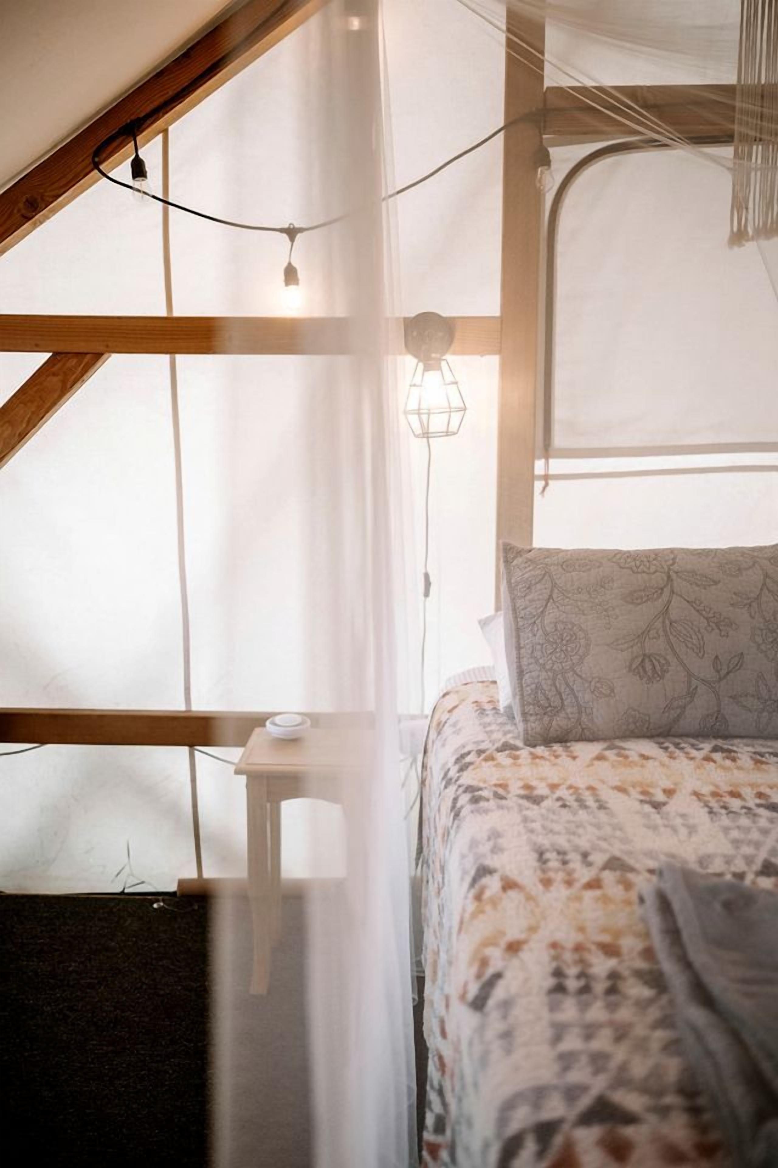 A bedroom with a bed, a small bedside table, a hanging light fixture, and a window with sheer curtains.