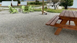 A gravel patio with a wooden picnic table and green chairs, a covered area, and a view of mountains and a lake.