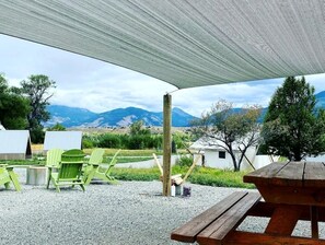 An outdoor seating area with a wooden picnic table and green Adirondack chairs, a wooden post, and a white tent in the background. - Stunning Safari Tent for Two near Yellowstone for a Memorable Glamping Escape in Emigrant, Montana (Emigrant)