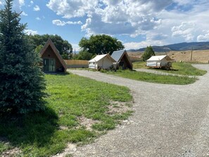 Un chemin de gravier mène à trois petites cabanes dotées de toits triangulaires, situées dans une zone herbeuse parsemée d’arbres, sous un ciel dégagé.