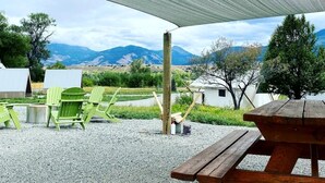 An outdoor seating area with a wooden picnic table and green Adirondack chairs, a wooden post, and a white tent in the background.