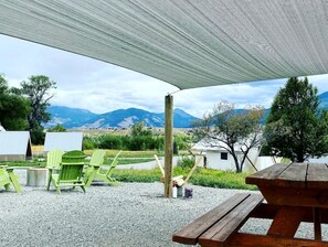 An outdoor seating area with a wooden picnic table and green Adirondack chairs, a wooden post, and a white tent in the background.