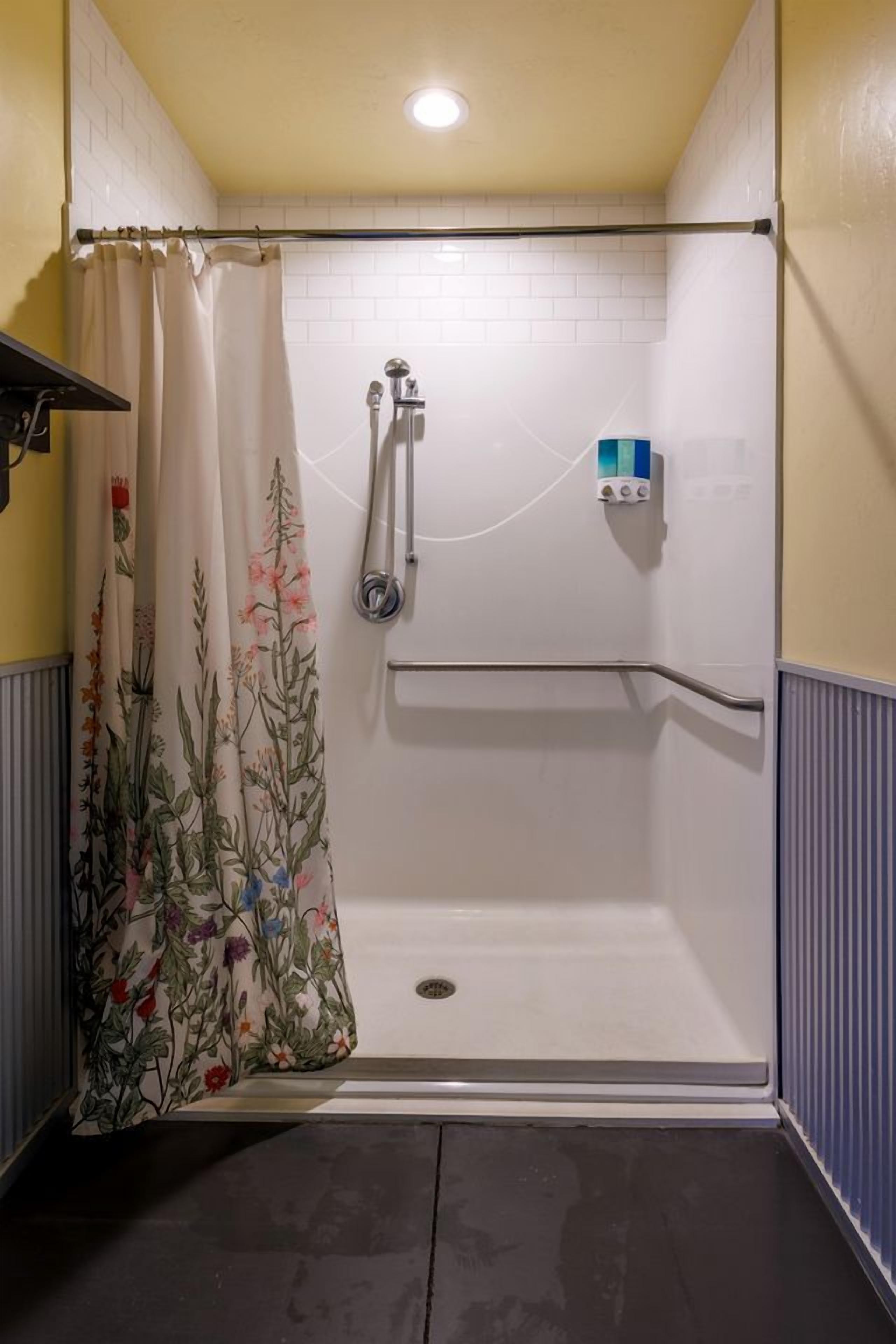 Une douche avec un mur recouvert de carreaux blancs, un rideau de douche à motifs floraux et une barre d’appui.