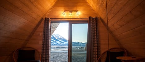 Ein Zimmer mit schrägen Wänden, einem Bett, zwei Korbstühlen, einem kleinen Tisch und Blick auf einen verschneiten Berg durch eine Glastür.