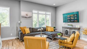 Una sala moderna con un sofá gris, sillones amarillos, una mesa de centro y un televisor montado en la pared mostrando un anuncio de inmueble.