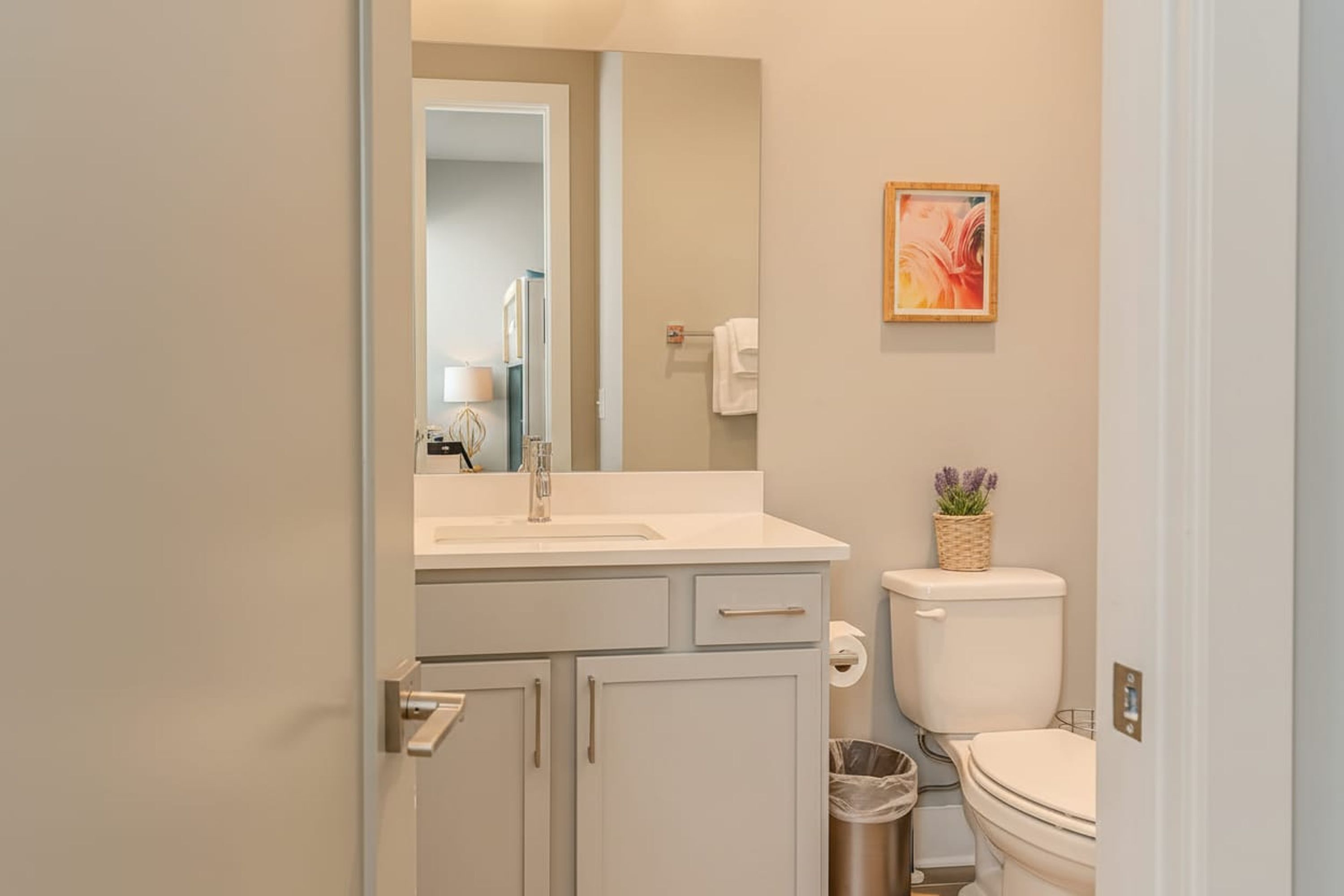 A bathroom with a white sink, mirror, and toilet. There is a trash bin and a small potted plant on the toilet.