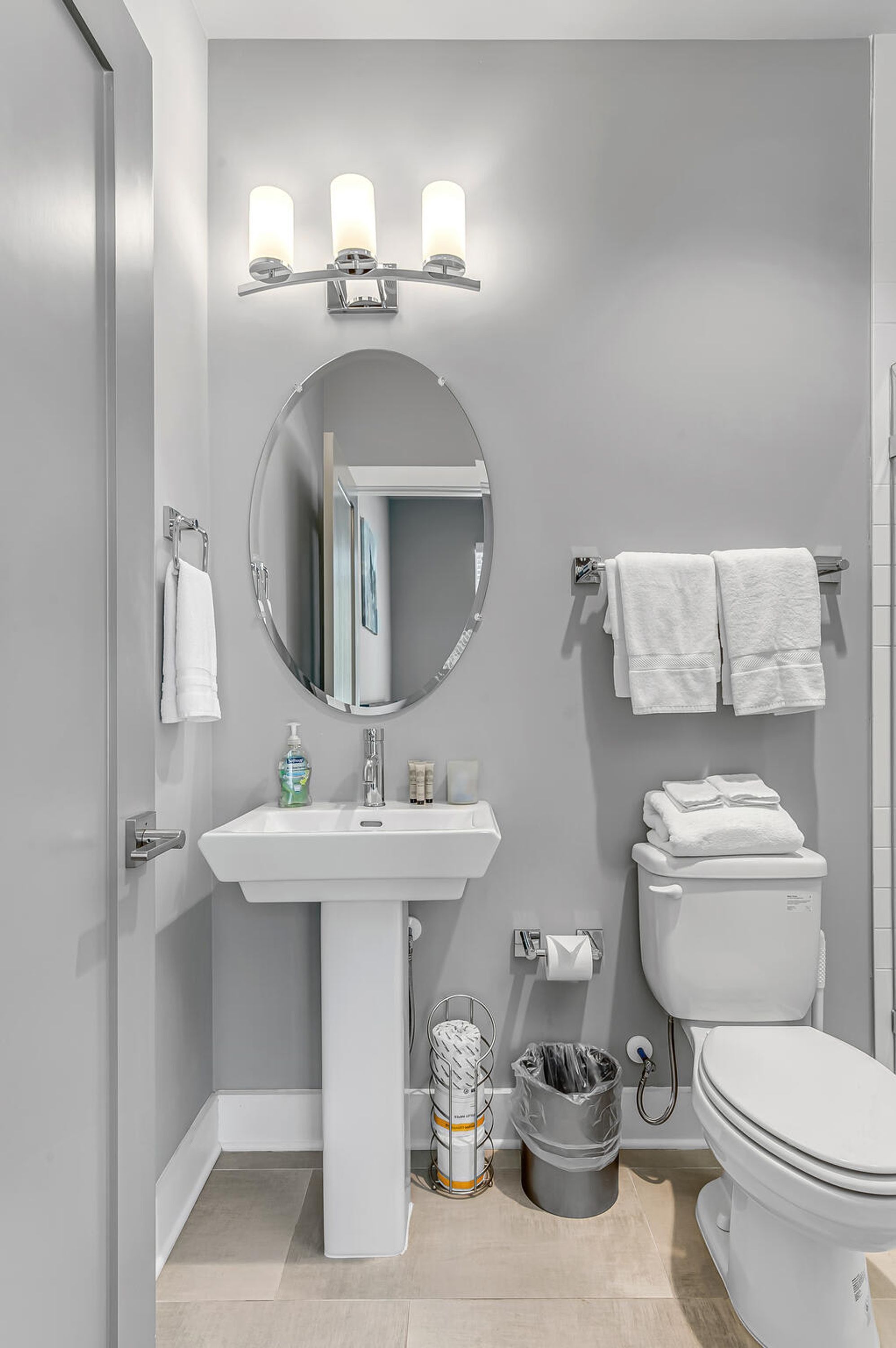 Un baño moderno con lavamanos blanco, espejo y retrete. Hay tres luces empotradas sobre el lavamanos y un perchero con toallas.