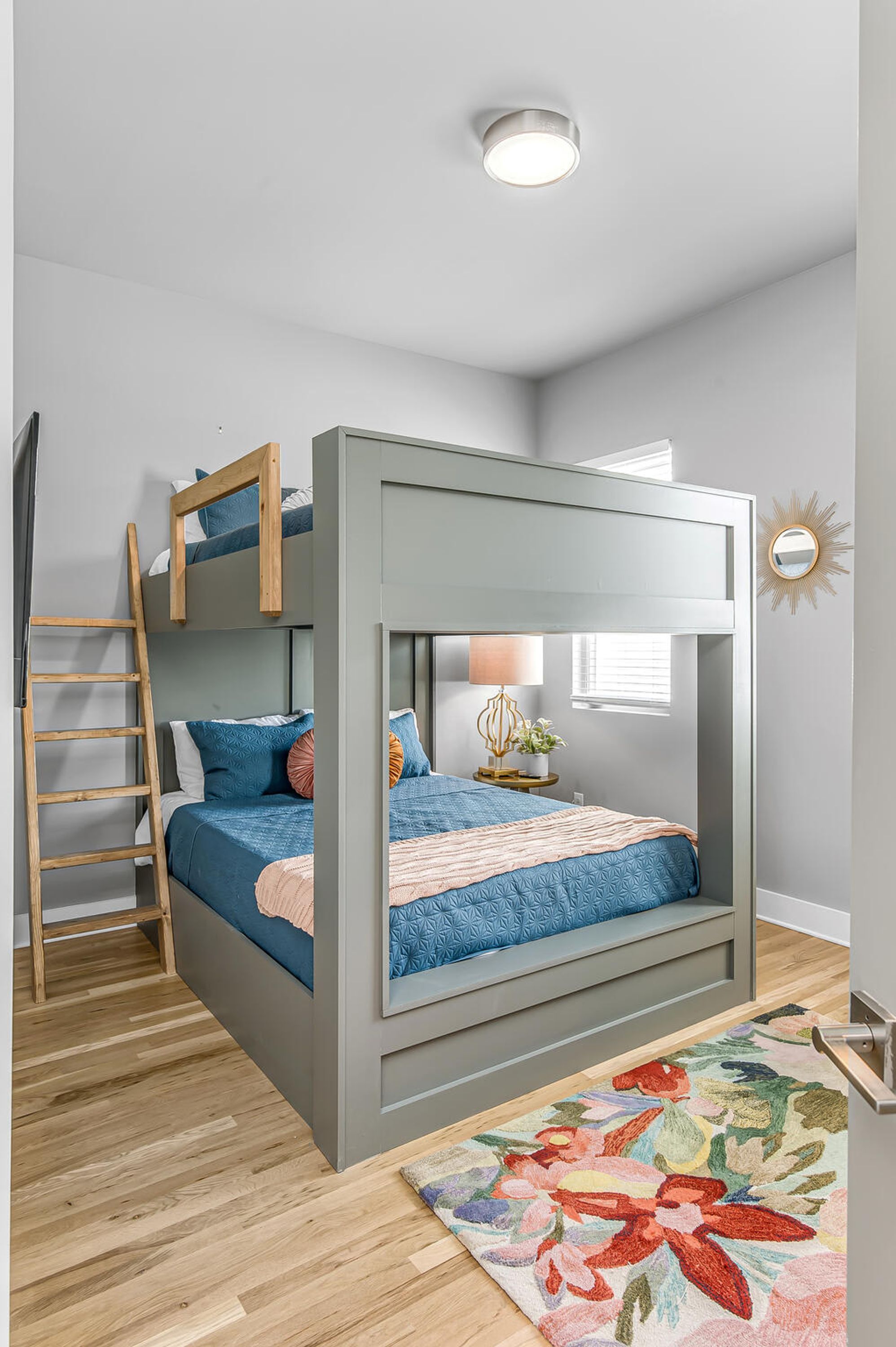A bunk bed with a ladder, a bedside table with a lamp, and a floral rug on a wooden floor.