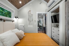 Une chambre à coucher comprenant un lit, des lampes de chevet, un climatiseur fixé au mur et donnant sur la cuisine adjacente.