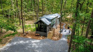 A cabin in a forest with a gravel driveway and wooden steps leading to a metal roof.