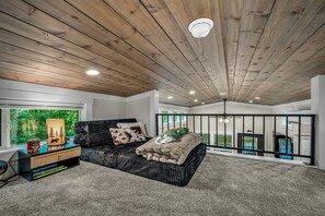 Une chambre avec un plafond en bois, un lit recouvert d’un plaid en fourrure, une table de chevet avec des livres et une fenêtre donnant sur un paysage verdoyant.