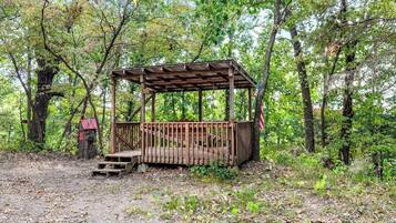 A wooden gazebo with a roof and a fenced area in a wooded setting.