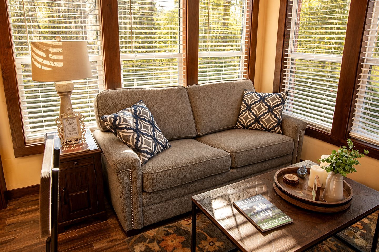 A living room with a sofa, two patterned pillows, a side table with a lamp, a coffee table with a book and decorative items, and a window with blinds.