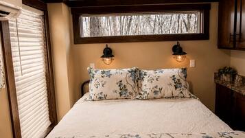 A bedroom with a bed, two wall-mounted lights, a window with blinds, and a view of trees.