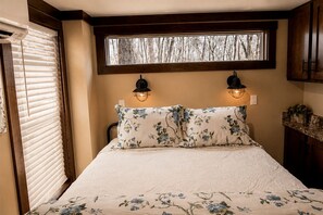 A bedroom with a bed, two wall-mounted lights, a window with blinds, and a view of trees. - Picturesque Tiny Cottage with Cozy Indoor and Outdoor in Tennessee (Monteagle)