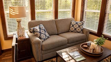 A living room with a sofa, two patterned pillows, a side table with a lamp, a coffee table with a book and decorative items, and a window with blinds.