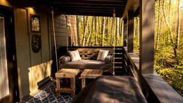 A covered porch with a wooden bench and a cushioned seating area, surrounded by trees.