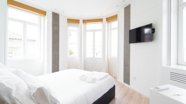 Ein modernes Schlafzimmer mit einem großen Bett, einem flachbildfernseher an der Wand und großen Fenstern mit gelben Jalousien.