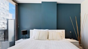 A modern bedroom with a large bed, a bedside table with a lamp, and a view of the city through a window. - Modern City Convenience With Great Amenities (Melbourne)