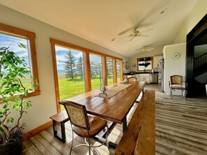 A spacious dining area with a long wooden table, chairs, and a view of a green landscape through large windows. - Spacious Farmhouse Estate with Mountain Views & Dual Kitchens near Skiing and Trails in Tetonia, Idaho (Tetonia)