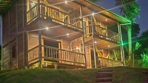 A two-story wooden house with a balcony, a stone staircase leading up to the entrance, and a palm tree in the background. - Chácara da Sorte (Pacoti)