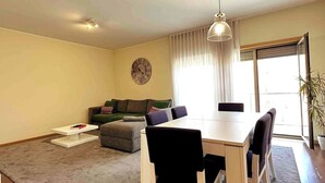 Un salon avec un canapé, une table à manger et des chaises, une horloge au mur et une grande fenêtre avec des rideaux.