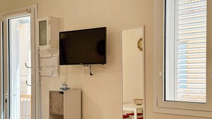 Una habitación con un televisor montado en la pared, un armario blanco, una pequeña mesa con un objeto decorativo y un espejo que refleja una silla y una mesa.
