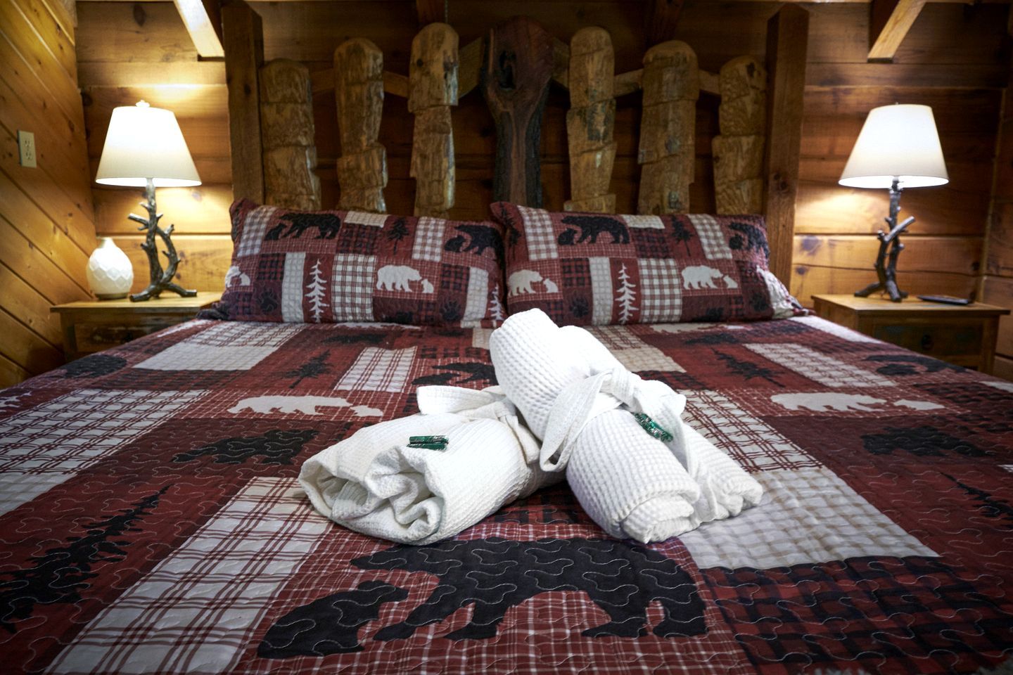 Una habitación tipo cabaña de madera con una cama, dos mesitas de noche y un cabecero con postes de madera tallada.