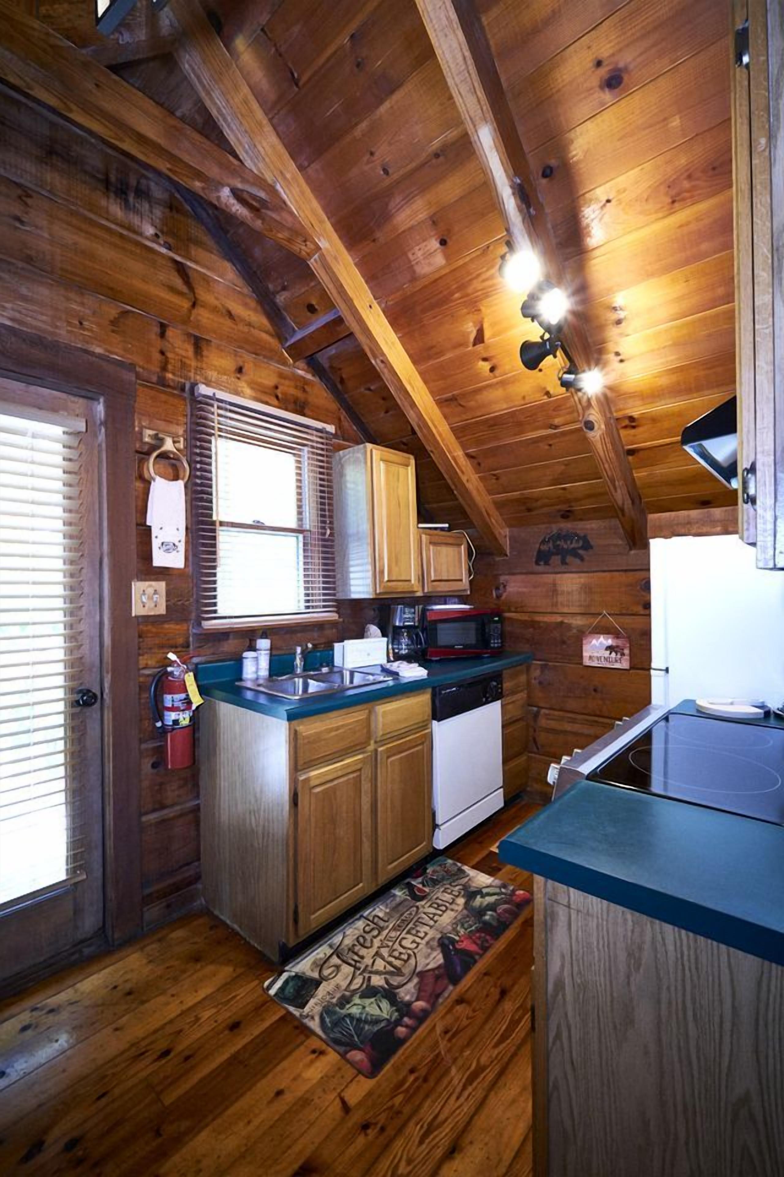 A wooden cabin interior with a kitchen area, wooden ceiling beams, and a window with blinds.