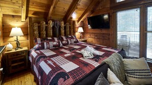 Une chambre à coucher de style cabane en bois, avec un grand lit, deux lampes de chevet, un téléviseur fixé au mur et une fenêtre avec des stores.