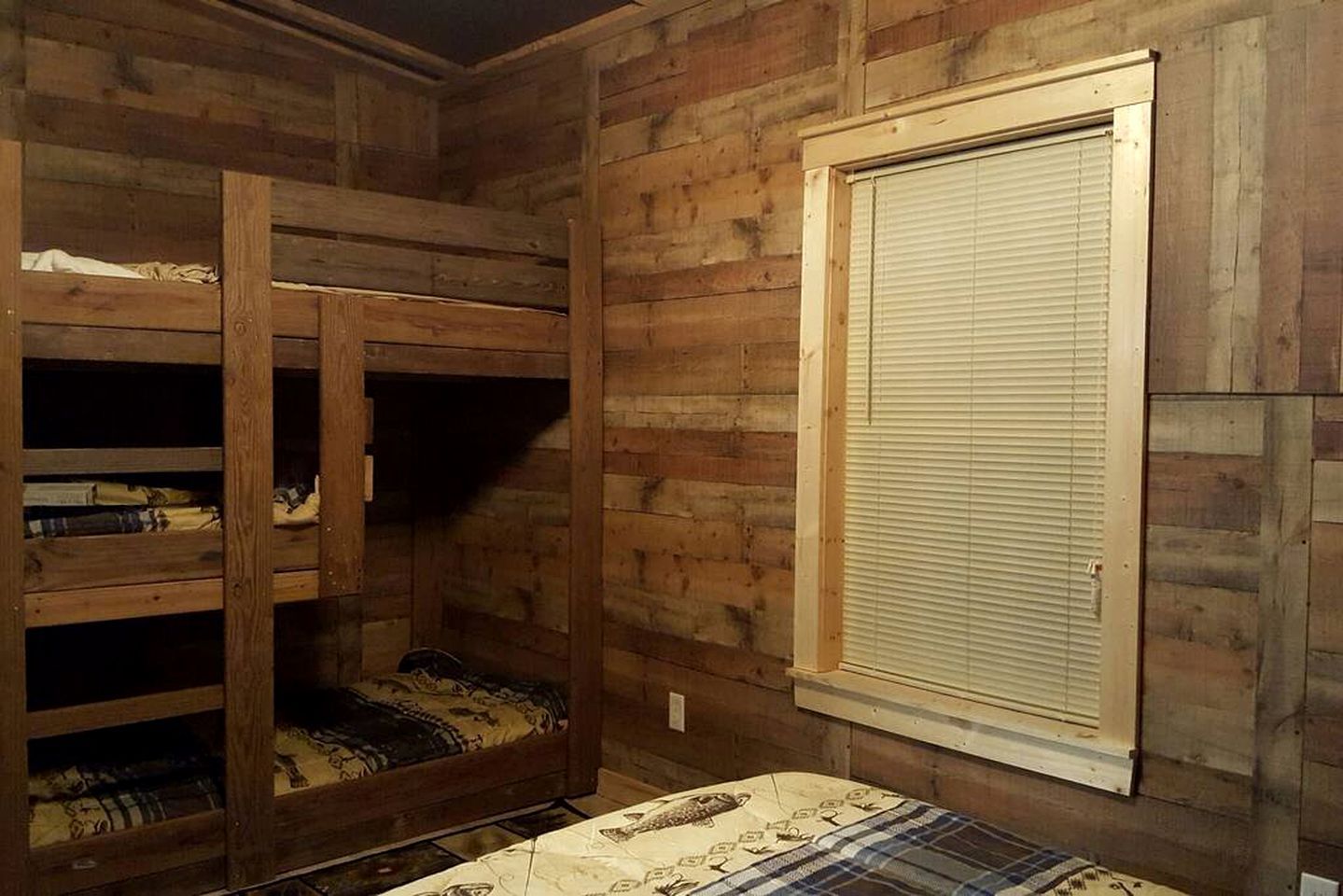 A room with wooden walls, a window with blinds, and a bunk bed with a patterned bedspread.