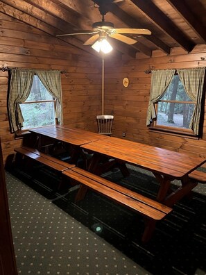 Ein Holzblockhaus-Interieur mit einem großen Esstisch, zwei Fenstern mit Karomustert-Vorhängen, einem Deckenventilator und einer Lampe.