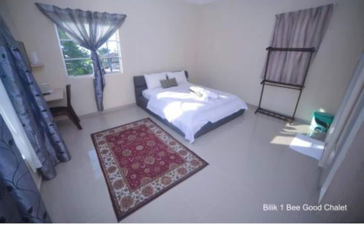 Ein Schlafzimmer mit Bett, Fenster mit Vorhängen, einem Sessel, einem kleinen Tisch und einem Wandregal.