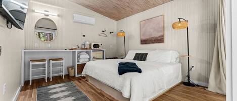 Une chambre à coucher avec un lit, une table de chevet et un bureau avec une chaise. Il y a une fenêtre, une lampe et un tableau au mur.